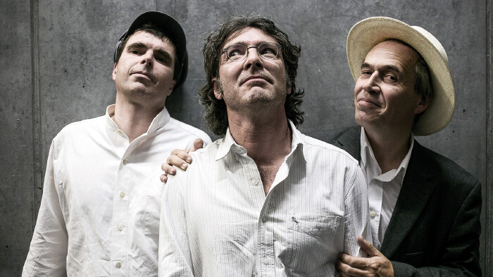 Drei Männer stehen dicht nebeneinander vor einer Betonwand. Der Mann links trägt ein weißes Hemd und eine dunkle Schirmmütze; er blickt leicht nach oben.