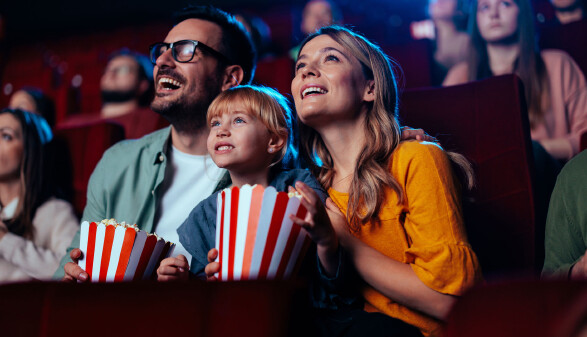 Eine Familie - Vater, Mutter, Tochter - sitzt mit 2 gro&szlig;en T&uuml;ten Popcorn im Kinosaal und hat sichtlich Freude am Film.