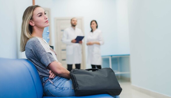 Junge Frau sitzt im Krankenhaus und wartet auf einen Arzttermin, im Hintergrund sprechen ein Arzt und eine &Auml;rztin miteinander