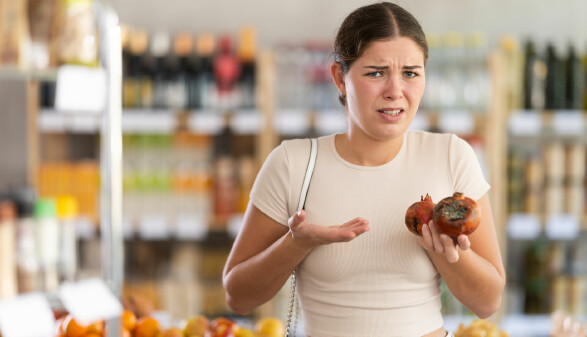 Eine junge Frau steht in einem Supermarkt vor einem Obstregal. Sie hält zwei verschimmelte Granatäpfel in den Händen und schaut verärgert und verwundert. Im Hintergrund sind weitere Obstsorten und Regale mit Lebensmitteln zu sehen.