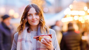 Eine junge Frau mit schulterlangen, gewellten Haaren steht im Freien und hält ein Brötchen mit einer langen Bratwurst und Ketchup in der Hand.