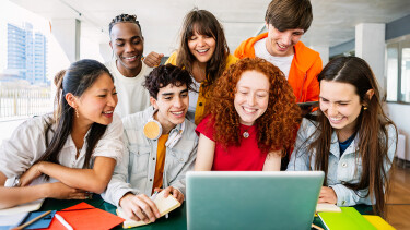 Eine fröhliche Gruppe junger Menschen sitzt gemeinsam vor einem Laptop und schaut lachend auf den Bildschirm.