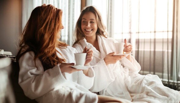 Zwei Frauen in Bademänteln trinken Kaffee oder Tee und lächeln sich in entspannter Atmosphäre an