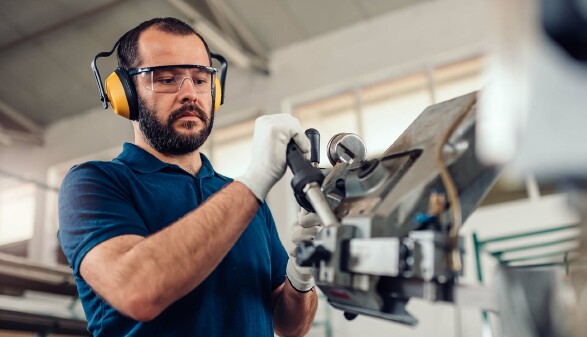 Ein Arbeiter in Schutzbrille, Geh&ouml;rschutz und Handschuhen bedient eine gro&szlig;e Metallmaschine in einer Werkhalle