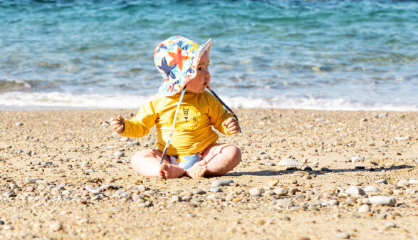 Ein Baby mit Badehut und UV-Shirt sitzt am Strand.