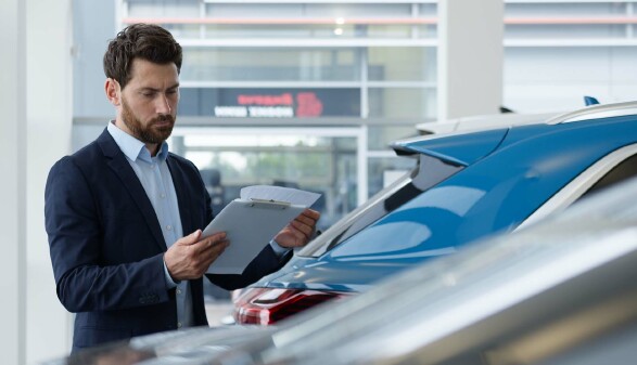 Ein Mann in Businesskleidung steht in einem Autohaus neben einem blauen Fahrzeug und prüft Unterlagen auf einem Klemmbrett.