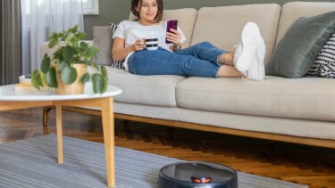 Ein Saugroboter vor einer Couch, auf der eine junge Frau liegt, eine Kaffeetasse in der Hand h&auml;lt und auf ihr Handy schaut