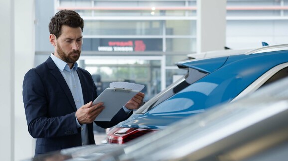 Ein Mann in Businesskleidung steht in einem Autohaus neben einem blauen Fahrzeug und prüft Unterlagen auf einem Klemmbrett.