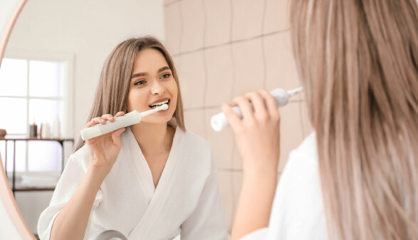 Junge Frau putzt sich die Z&auml;hne im Badezimmer und schaut sich dabei im Spiegel zu.