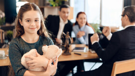 Junges M&auml;dchen, dass Teddyb&auml;r umarmt, im Hintergrund Eltern mit Berater