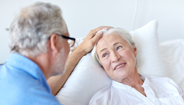 Pflegebedürftige Frau liegt im Bett und ihr Mann legt seine Hand auf ihren Kopf © SydaProductions, stock.adobe.com