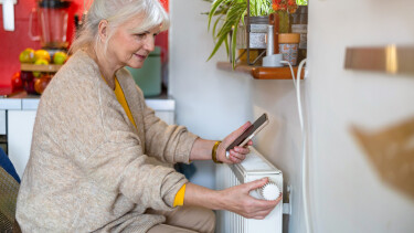 &Auml;ltere Frau h&auml;lt ein Handy in der Hand, w&auml;hrend sie das Thermostat am Heizk&ouml;rper zu Hause einstellt