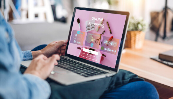 Person sitzt auf dem Sofa und nutzt einen Laptop, auf dem eine Online-Shopping-Seite mit Kosmetikartikeln und Anmeldefeldern angezeigt wird.