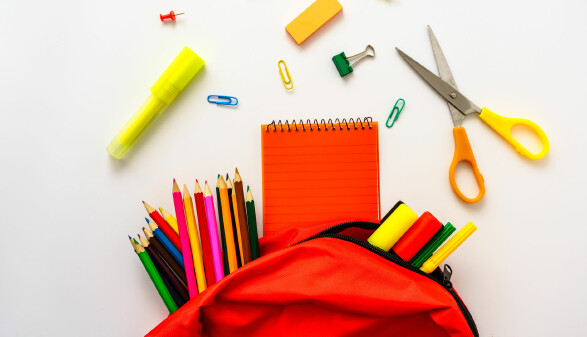 Bunte Schulmaterialien auf weißem Hintergrund, darunter Stifte, Notizbuch, Schere und ein roter Rucksack.