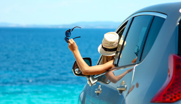 Junge Frau mit Strohhut und Sonnenbrille in der Hand schaut vom Auto aus aufs Meer.