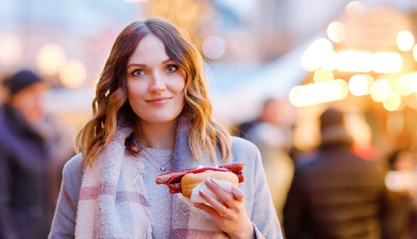 Eine junge Frau mit schulterlangen, gewellten Haaren steht im Freien und hält ein Brötchen mit einer langen Bratwurst und Ketchup in der Hand.