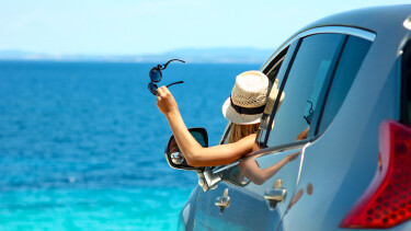 Junge Frau mit Strohhut und Sonnenbrille in der Hand schaut vom Auto aus aufs Meer.
