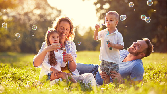 Eine Familie sitzt auf einer Wiese im Sonnenschein und spielt mit Seifenblasen; zwei Kinder pusten Seifenblasen, während die Eltern lächelnd daneben sitzen. © luckybusiness_low, stock.adobe.com Eine Familie sitzt auf einer Wiese im Sonnenschein und spielt mit Seifenblasen; zwei Kinder pusten Seifenblasen, während die Eltern lächelnd daneben sitzen.