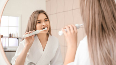 Junge Frau putzt sich die Z&auml;hne im Badezimmer und schaut sich dabei im Spiegel zu.