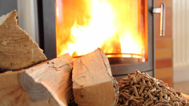 Ein brennender Kaminofen mit sichtbarer Flamme. Davor liegen Holzscheite und Holzpellets.
