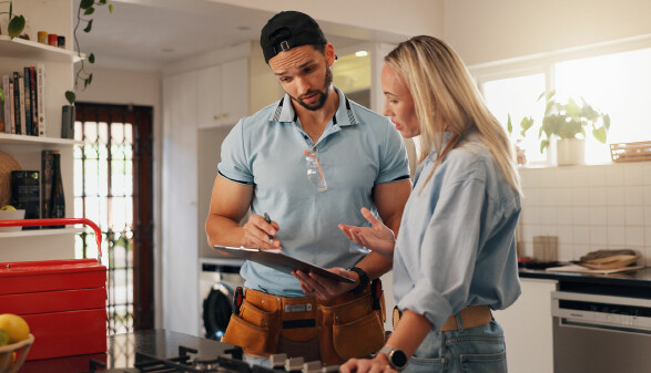 Ein Installateur und eine Frau stehen in einer hellen Küche und besprechen etwas auf einem Klemmbrett.