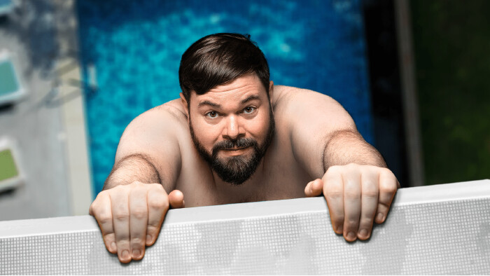 Ein oberkörperfreier Mann hält sich an einer Kante fest, hinter ihm ein blaues Schwimmbecken.