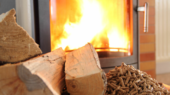 Ein brennender Kaminofen mit sichtbarer Flamme. Davor liegen Holzscheite und Holzpellets.