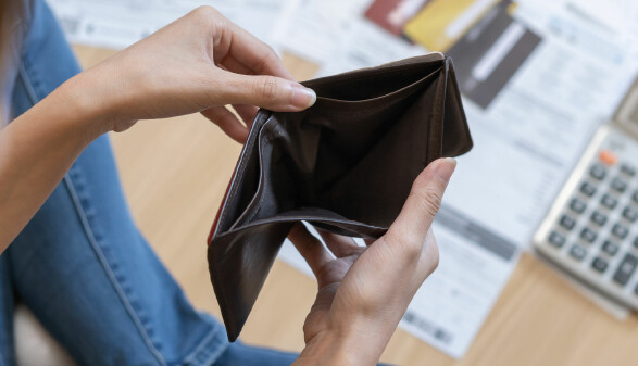 Eine Person hält eine leere Geldbörse in den Händen. Im Hintergrund liegen Rechnungen, Kreditkarten und ein Taschenrechner auf dem Tisch.